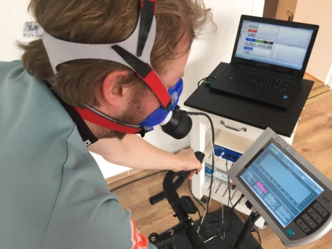 Ein Sportler mit Atemmaske und Stirnband beugt sich über den Lenker, im Hintergrund ein Laptop mit Messkurve