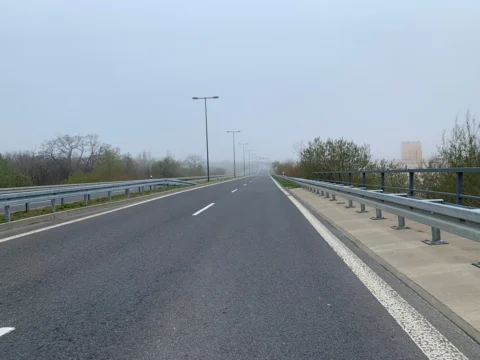 Eine leere Schnellstraße mit Leitplanke und Laternenreihe, am Horizont ein Gebäude