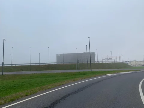 Eine leere Straße vor einer Wiese, dahinter Hallen und Masten im Dunst
