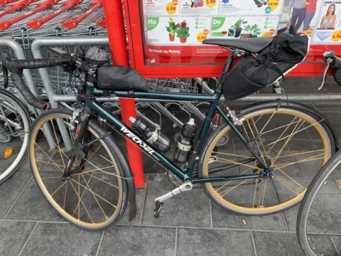 Ein grünes Reiserad mit Lenker- und Satteltasche am Fahrradständer vor einem Supermarkt
