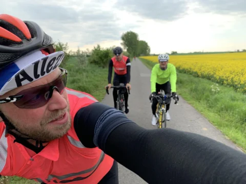 Selfie eines Fahrers, dahinter zwei Begleiter auf einem Weg zwischen Rapsfeldern