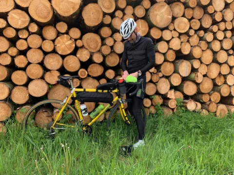 Ein Fahrer steht mit gelbem Rennrad vor einem hoch aufgeschichteten Holzstapel