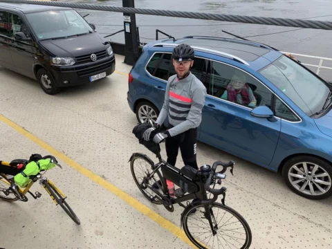 Ein Fahrer mit bepacktem Rennrad wartet an einer Ampel zwischen Autos
