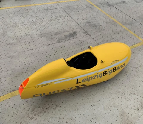 Ein gelbes Velomobil mit der Aufschrift „LeipzigBigBand“ steht auf einem Parkplatz