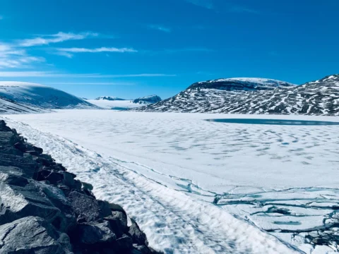 Ein noch zugefrorener Stausee unter blauem Himmel, ringsum schneebedeckte Hänge