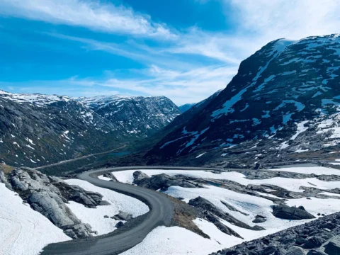 Eine Passstraße windet sich durch ein Tal mit Altschnee, dahinter dunkle Bergflanken