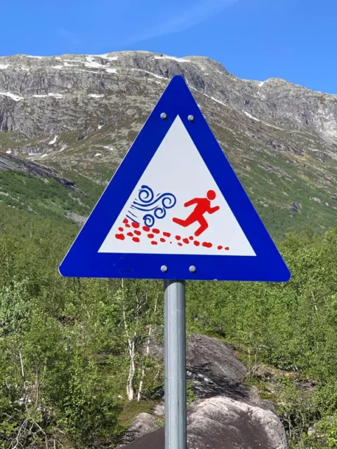 Ein blaues Warnschild mit einer rennenden Figur vor Wasser und Steinen, dahinter Berghänge