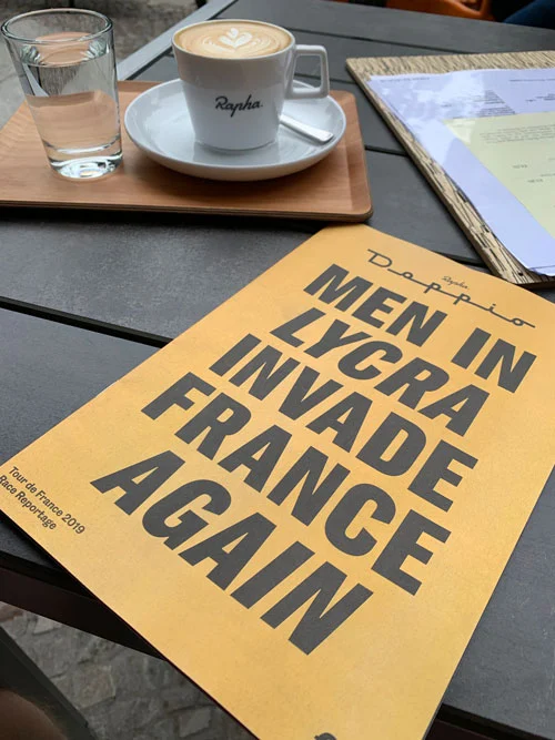 Ein Cappuccino auf einem Tisch neben einem Heft mit der Aufschrift „Men in Lycra invade France again“