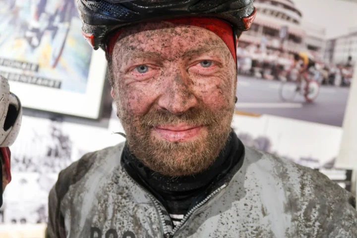 Ein Radfahrer mit über und über verschlammtem Gesicht und Trikot lächelt in die Kamera