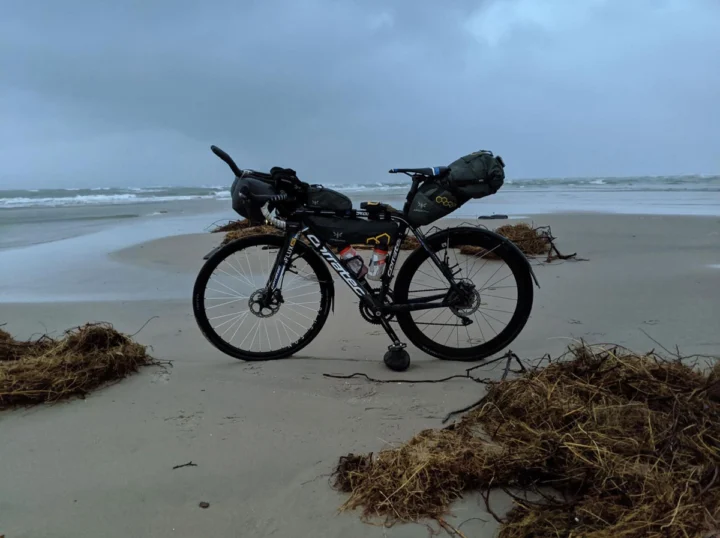Ein bepacktes Bikepacking-Rad steht am Strand zwischen Tanghaufen, dahinter das Meer