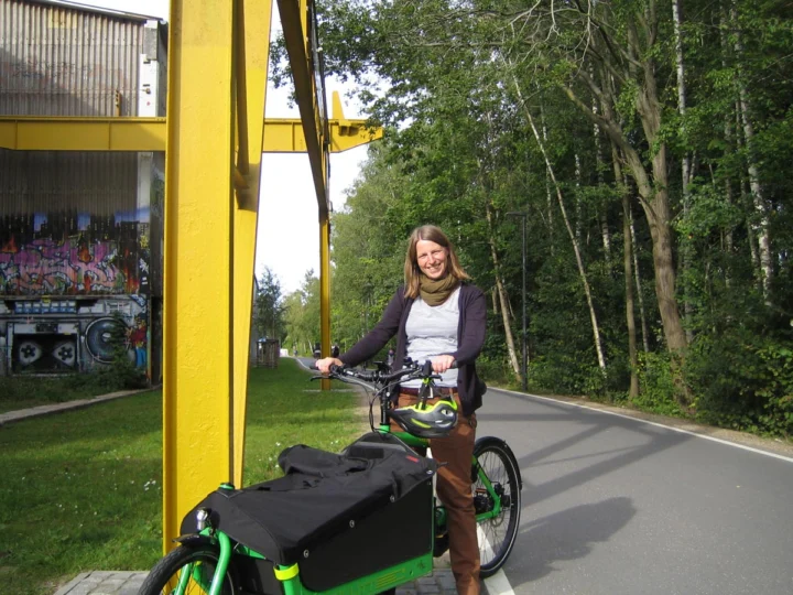 Eine Frau steht mit einem grünen Lastenrad auf einem Radweg unter einer gelben Brücke