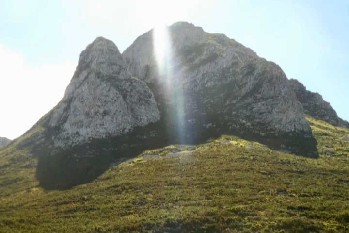 Ein zerklüfteter Felsgipfel über einem Grashang, dahinter die Sonne im Dunst