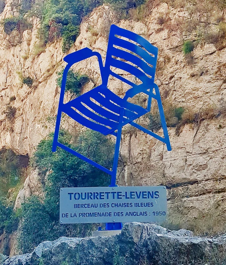 Ein überlebensgroßer blauer Stuhl an einem Felsen, darunter ein Schild „Tourrette-Levens, Wiege der Stühle der Promenade des Anglais“