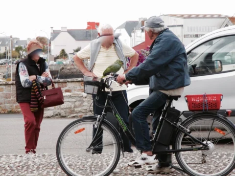 Zwei ältere Männer im Gespräch neben einem Fahrrad, auf dem Lenker sitzt ein grüner Papagei