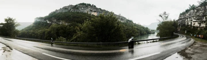 Eine nasse Straße am Fluss entlang, dahinter ein bewaldeter Felsrücken im Regendunst