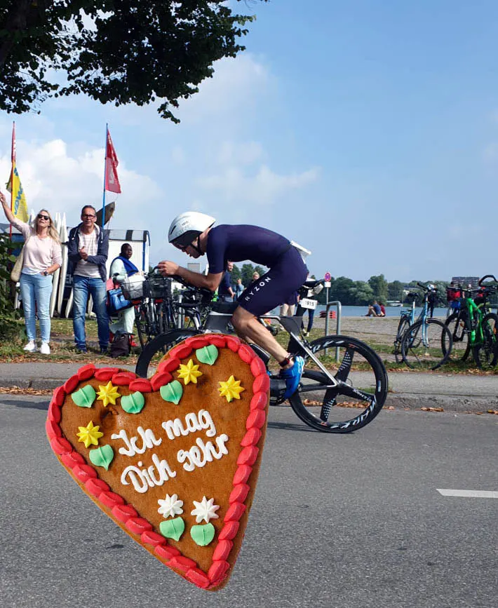 Ein Triathlet auf dem Zeitfahrrad am Seeufer, im Vordergrund ein Lebkuchenherz mit der Aufschrift „Ich mag Dich sehr“