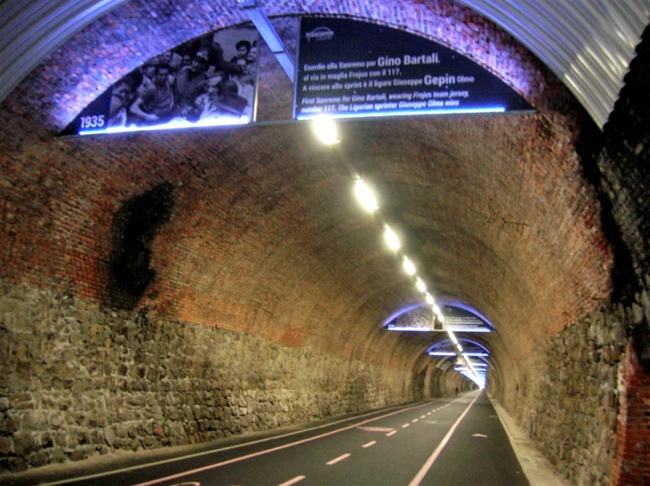 Ein beleuchteter Radweg durch einen alten Backsteintunnel, an der Decke Tafeln mit einem Foto von Gino Bartali