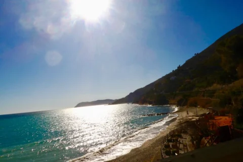 Die ligurische Küste im Gegenlicht, die Sonne glitzert auf dem Meer, rechts fällt ein steiler Hang zum Kiesstrand ab