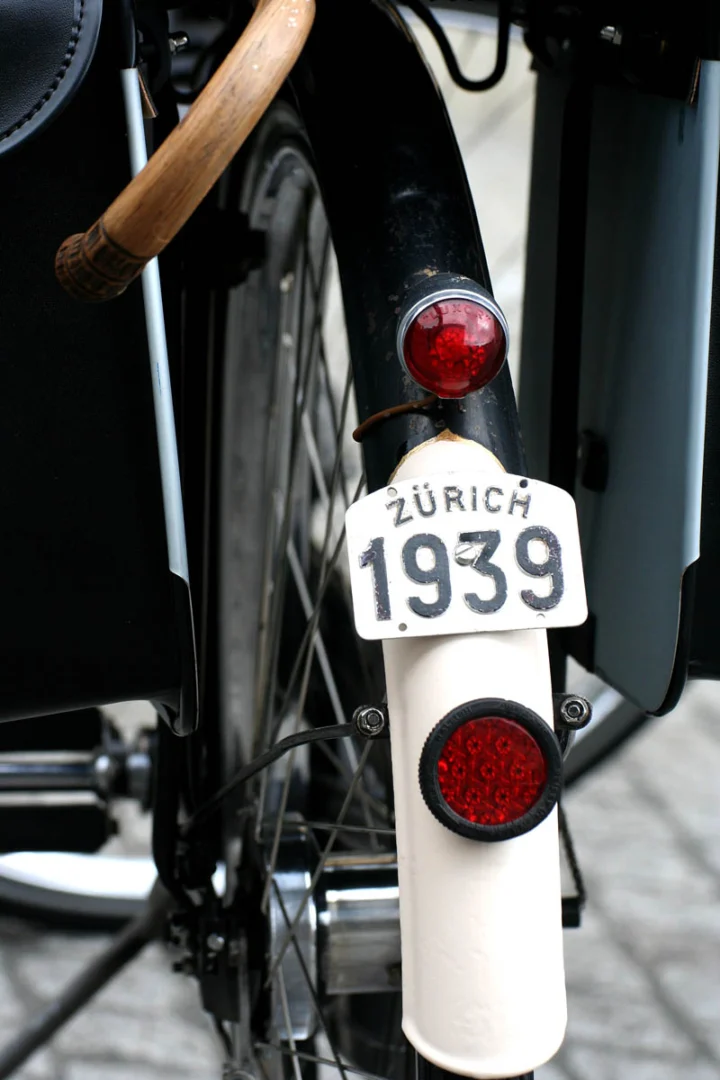 Ein schwarzes Fahrrad von hinten, am Schutzblech ein weißes Nummernschild „Zürich 1939“ über dem Rückstrahler