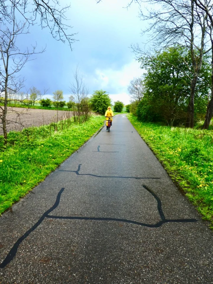 Ein Kind im gelben Regenanzug radelt einen schmalen Radweg zwischen Feldern entlang, am Himmel eine dunkle Regenwand