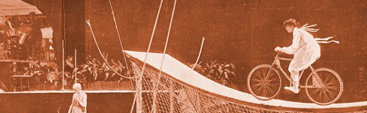 Historische Aufnahme in Sepia: eine Artistin fährt mit dem Fahrrad eine steile Rampe hinab