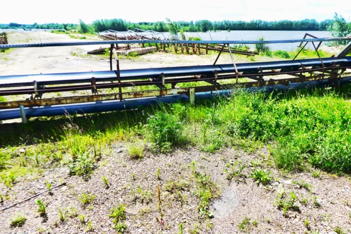 Förderbänder eines Kieswerks am Wasser, davor sandiger Boden mit Grasbüscheln