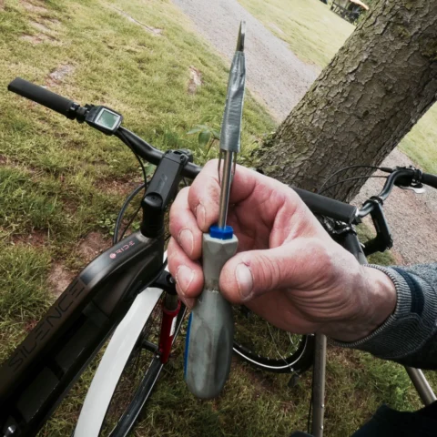 Hand hält einen Schraubendreher, in dessen Klinge eine Pinzette steckt, dahinter ein Fahrradlenker