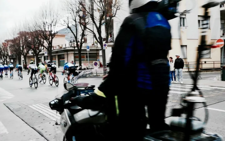 Ein Fahrerfeld biegt um eine Straßenecke, im unscharfen Vordergrund ein Zuschauer mit Fahrrad