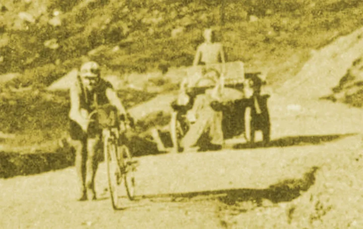 Historische Aufnahme in Sepia: ein Fahrer schiebt sein Rad einen Schotterpass hinauf, daneben Zuschauer