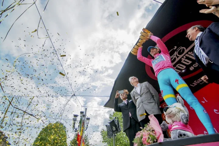 Ein Fahrer im Rosa Trikot fährt durch Konfettiregen, dahinter das Giro-Podium