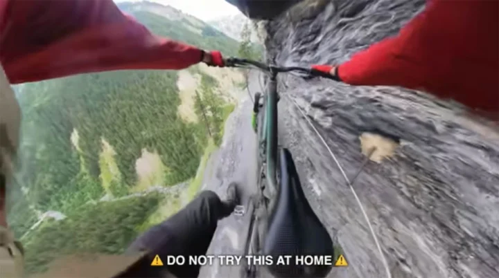 Bildschirmfoto eines Videos: Blick vom Lenker über eine Felskante steil ins Tal, eingeblendet die Warnung „Do not try this at home“