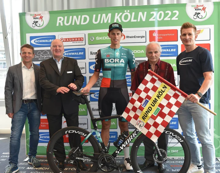 Pressekonferenz zu Rund um Köln 2022: fünf Personen vor der Sponsorenwand, in der Mitte der Sieger im Bora-Trikot mit der Zielflagge