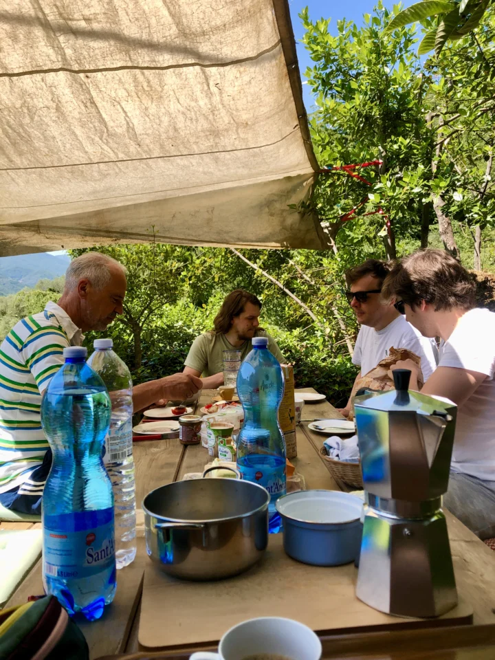 Das at-Team beim Frühstück unter einem Sonnensegel: Jens, Jonathan, Fabian und Daniel, dahinter Olivenbäume und Berge