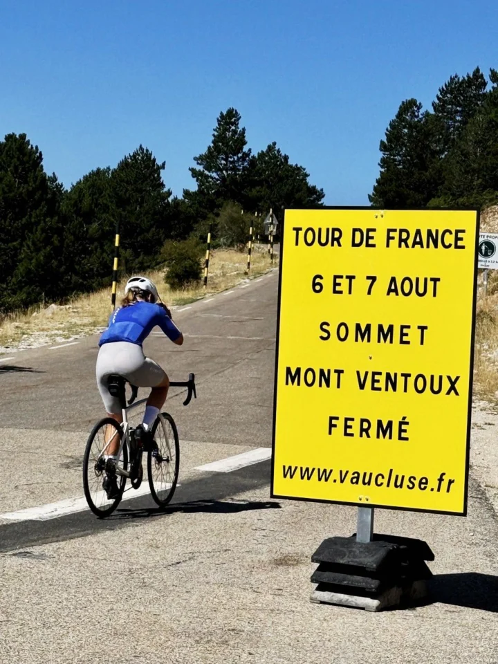 Eine Rennradfahrerin im blauen Trikot fährt an einem gelben Schild vorbei: „Tour de France 6 et 7 août, Sommet Mont Ventoux fermé“